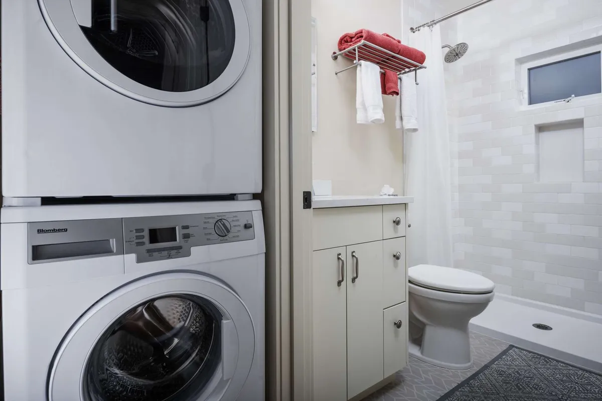 A practical interior showing an in-unit washer and dryer tucked beside the bathroom's sink and toilet, presented in a clean and organized arrangement. The space feels efficient and user-friendly, supporting self-care and daily convenience.