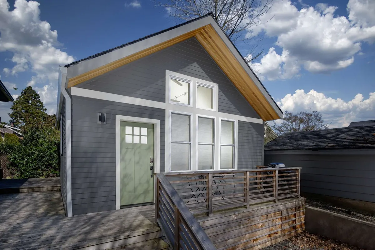 This exterior shot captures the front of the detached cottage, highlighting the green front door, distinctive angled roofline, and large windows. The image also reveals the front deck area, with glimpses of outdoor seating and a welcoming atmosphere. The space feels both private and inviting, suggesting a spot perfect for morning coffee, relaxing evenings, or hosting small gatherings.