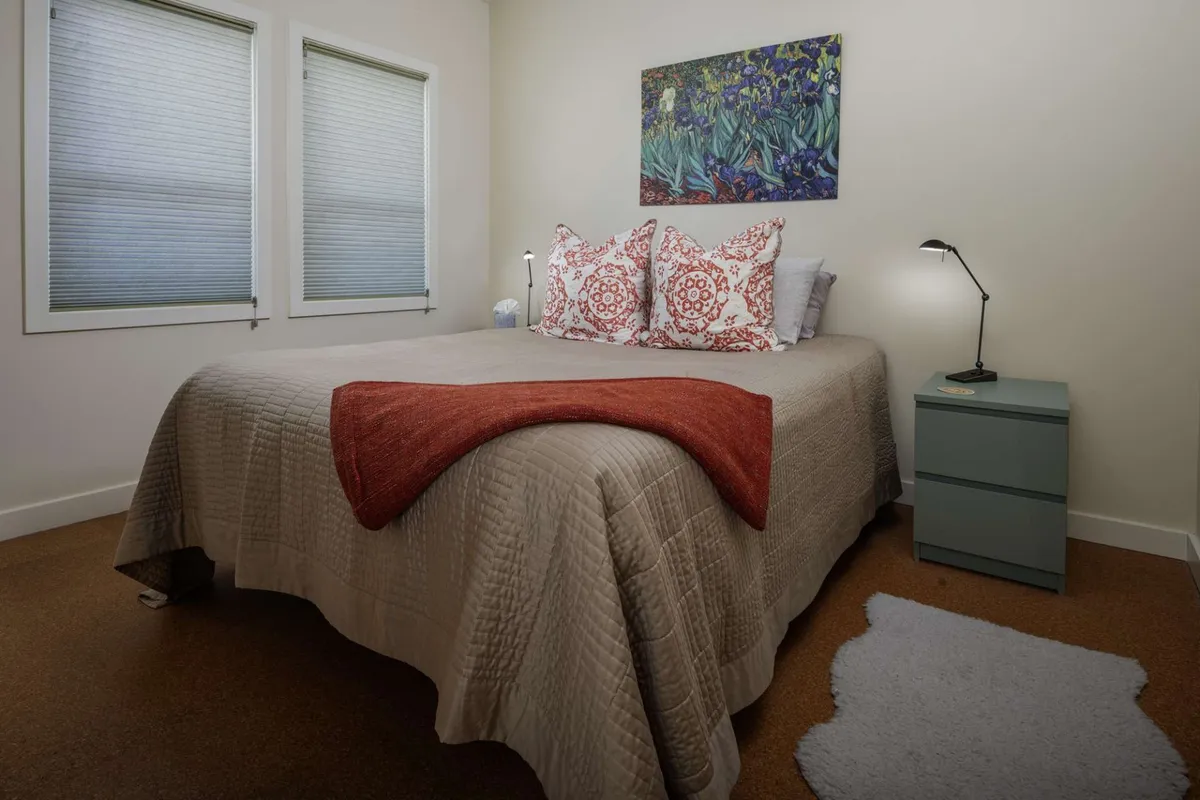 This image captures a cozy, inviting bedroom with a queen bed adorned with decorative pillows. The nightstand sits close by, illuminated by a reading light, while a calming painting hangs above the headboard, creating a peaceful ambiance ideal for rest and privacy.