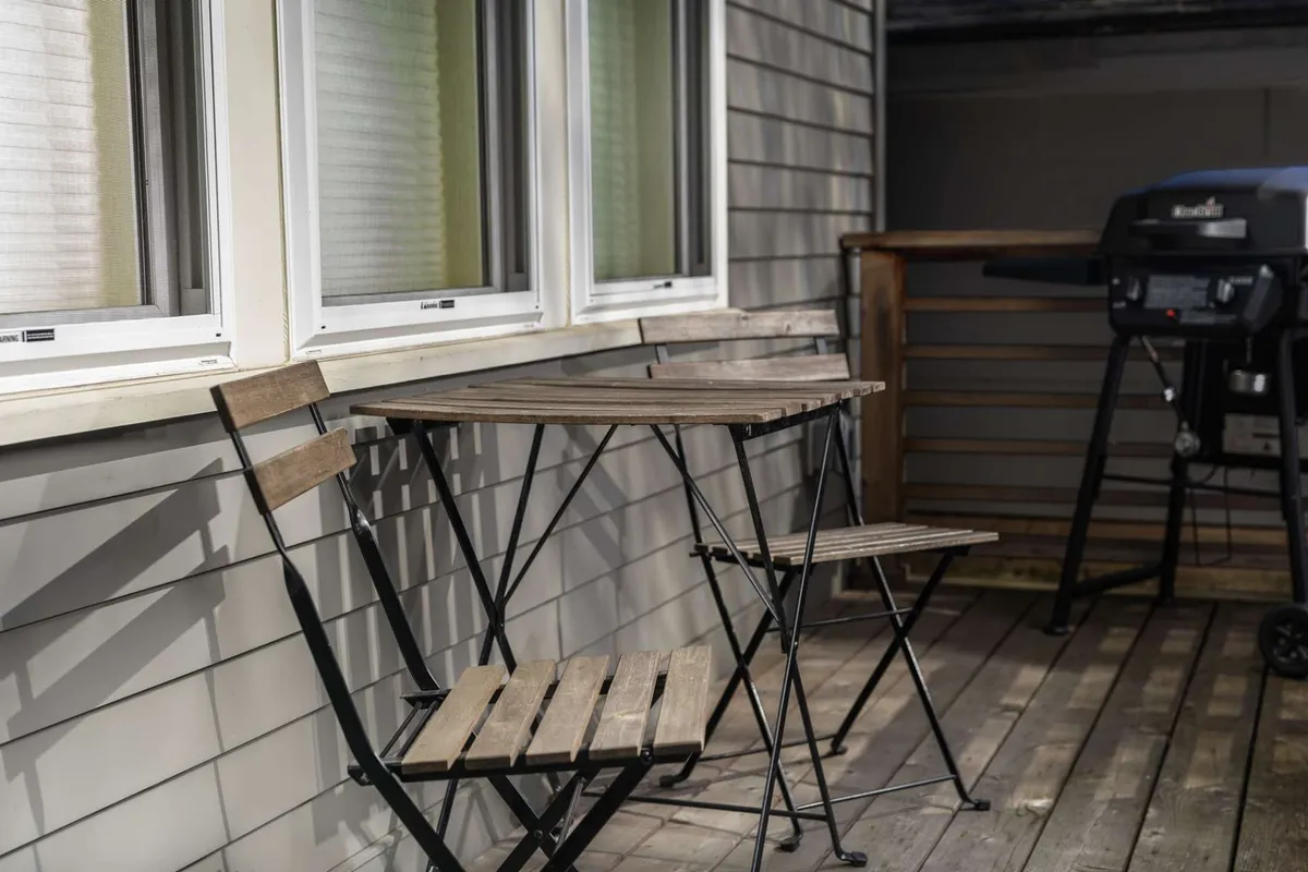 A close-up of the outdoor living space, featuring a wooden table, two folding chairs, and a BBQ grill. The setup suggests relaxed outdoor meals or quiet moments with a book. The lighting hints at either early morning or late afternoon, evoking both sunrise breakfasts and sunset barbecues as described in the section.